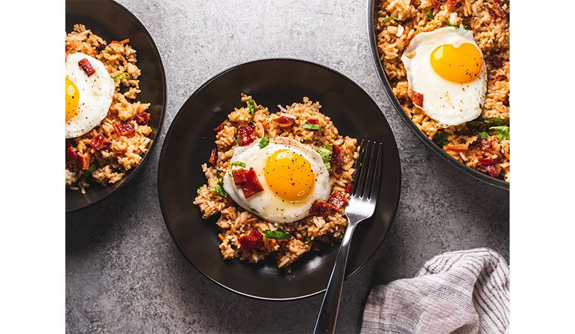 Three black bowls filled with fried rice, vegetables, and topped with sunny-side-up eggs, with a fork in one bowl, placed on a gray surface next to a striped cloth.