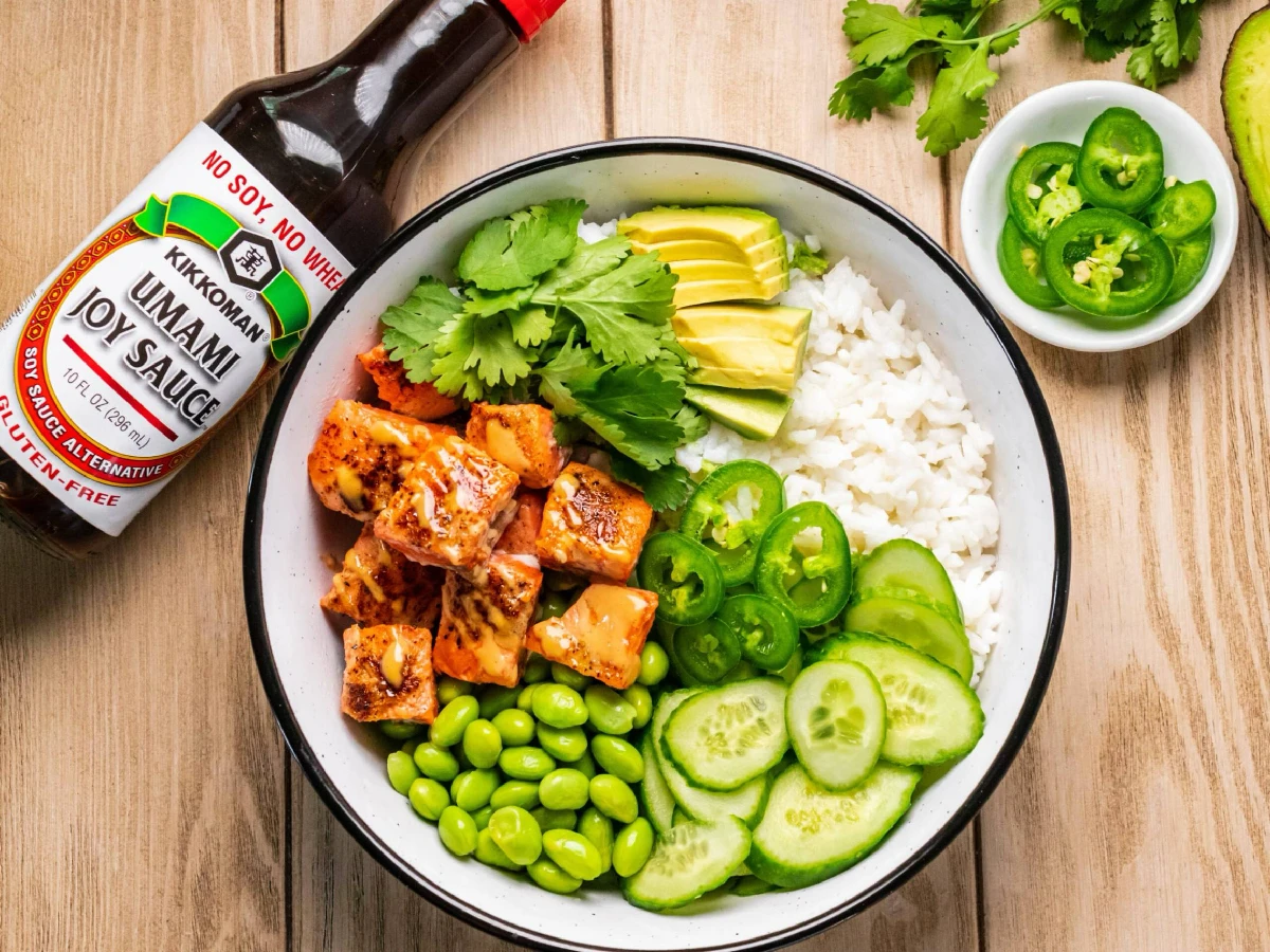 A bowl with white rice, grilled salmon cubes, edamame, sliced cucumber, avocado, cilantro, and jalapeños, with a bottle of Kikkoman Umami Soy Sauce and extra jalapeños on the side.