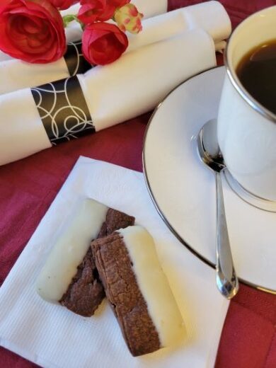 Two chocolate-dipped cookies on a napkin next to a cup of coffee with a spoon, white napkins, and red artificial roses on a red tablecloth.