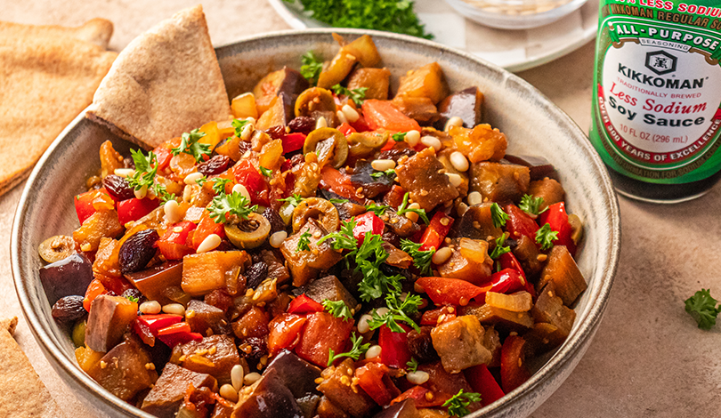 A bowl of caponata with eggplant, red peppers, olives, pine nuts, and herbs, served with pita bread; a bottle of soy sauce is in the background.