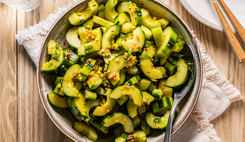 A bowl of sliced cucumber salad garnished with chopped garlic and spices, served with a spoon on a wooden table.