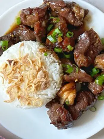 A plate with white rice topped with fried shallots and pieces of glazed grilled beef garnished with chopped green onions.