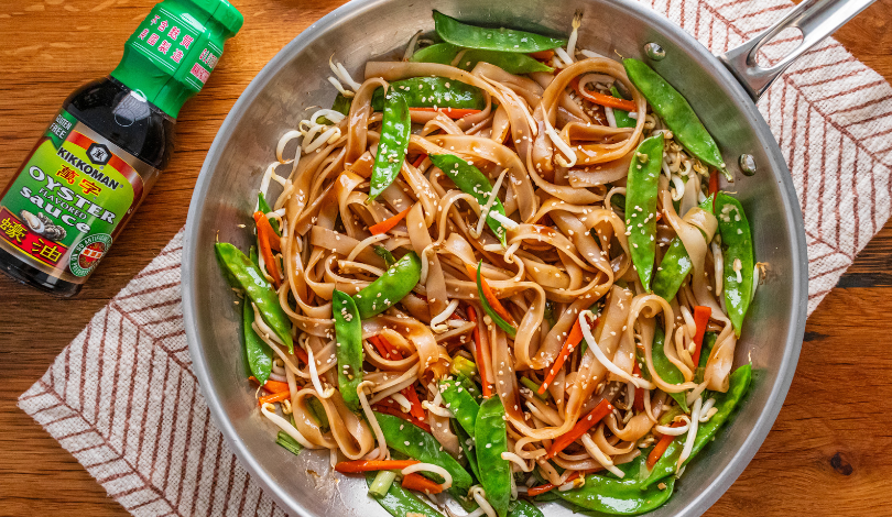 A pan of stir-fried noodles with snap peas, carrots, and bean sprouts, topped with sesame seeds, sits on a towel next to a bottle of Kikkoman oyster sauce.