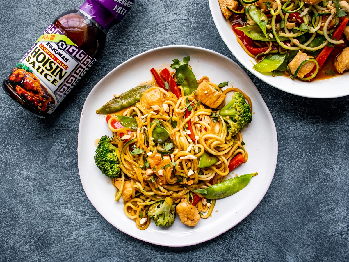 A plate of stir-fried noodles with chicken, broccoli, snow peas, and red peppers next to a bottle of Kikkoman hoisin sauce on a dark surface.