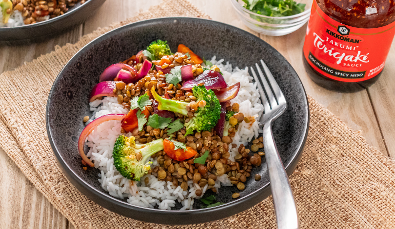 A bowl of white rice topped with broccoli, red onion, red pepper, lentils, and teriyaki sauce, with a fork on the side. A bottle of teriyaki sauce is visible in the background.