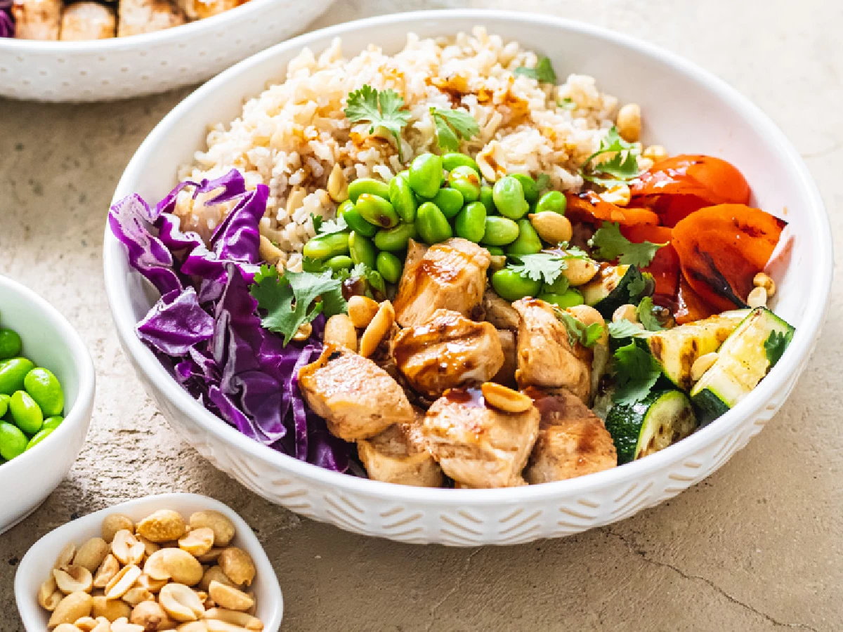 A white bowl filled with brown rice, grilled chicken, edamame, red cabbage, tomatoes, zucchini, peanuts, and cilantro.