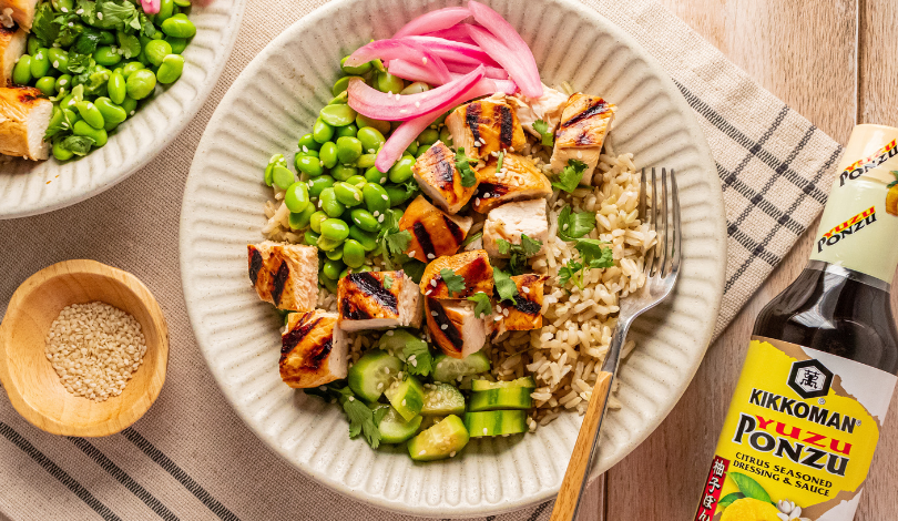A bowl of brown rice topped with grilled chicken, edamame, cucumbers, pickled onions, and cilantro, with a bottle of Kikkoman Yuzu Ponzu sauce nearby.