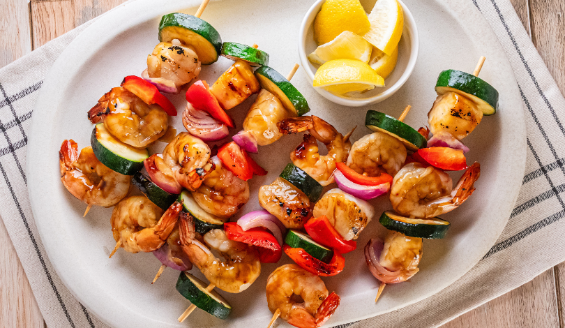 Grilled shrimp skewers with zucchini, red onion, and red bell pepper on a white plate, served with a small bowl of lemon wedges.