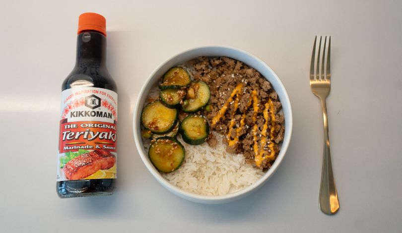 A bowl of white rice topped with seasoned ground meat, sliced cucumbers, and sauce, next to a bottle of Kikkoman Teriyaki sauce and a fork on a white surface.