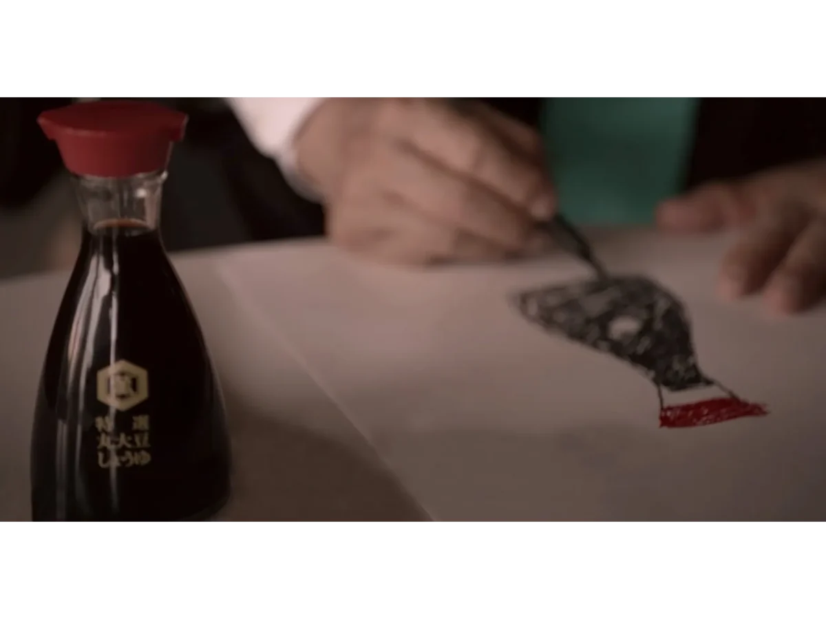 A soy sauce bottle sits on a table while a person uses a brush to paint a black and red shape on white paper in the background.