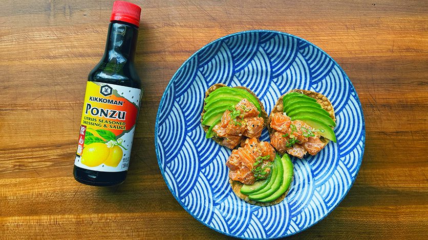 A bottle of Kikkoman Ponzu sauce sits beside a blue patterned plate with three crackers topped with diced salmon and sliced avocado on a wooden surface.