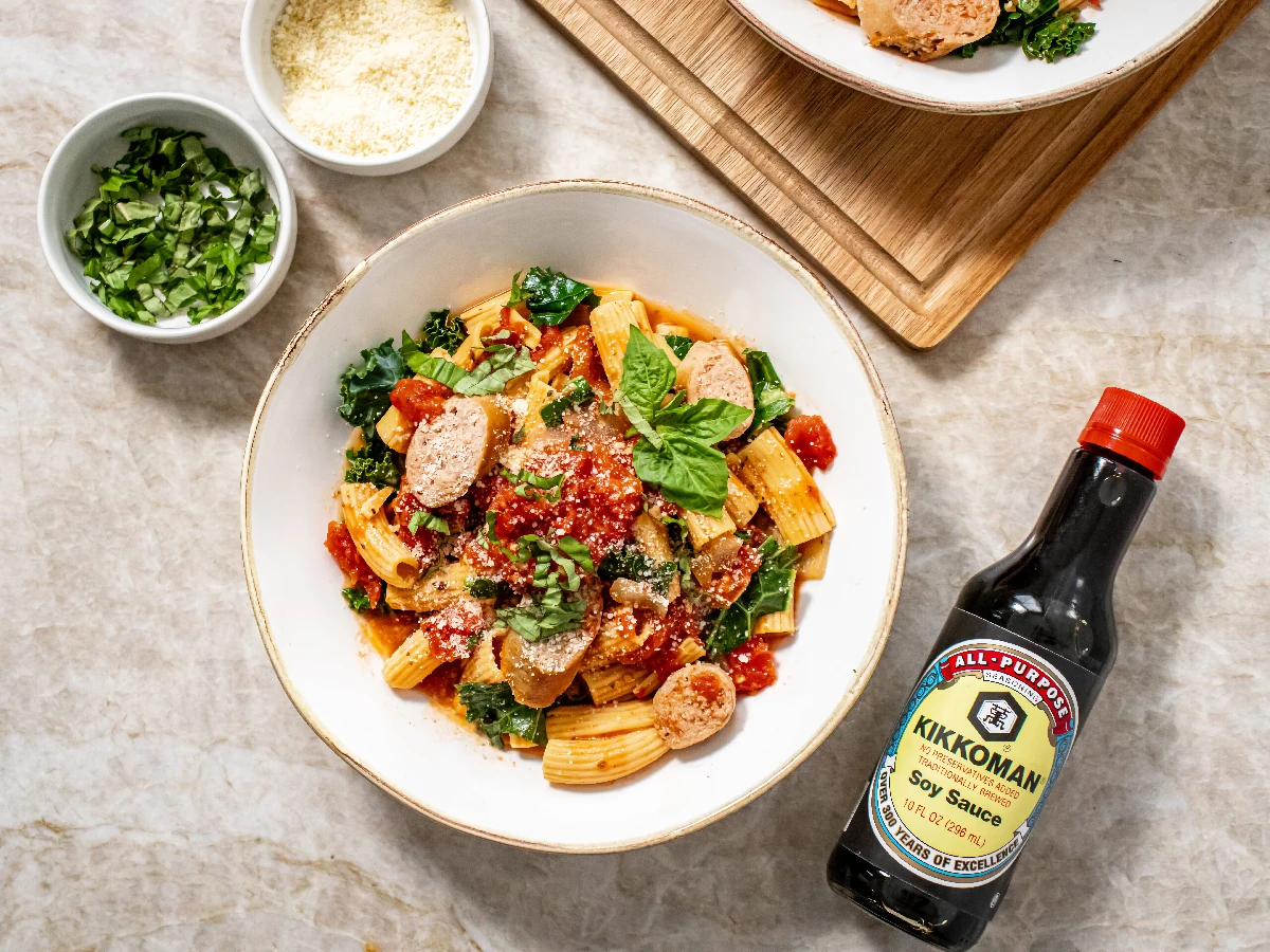 A bowl of rigatoni pasta with meatballs, tomato sauce, greens, and basil, next to a bottle of Kikkoman soy sauce, shredded cheese, and chopped herbs.