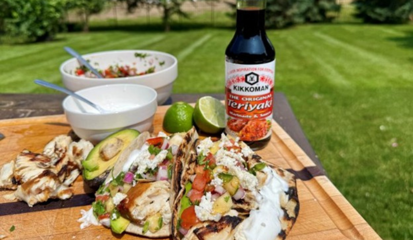 Grilled chicken tacos with avocado, salsa, and cheese on a wooden board, with lime wedges, salad bowls, and a bottle of Kikkoman Teriyaki Sauce on a picnic table outdoors.