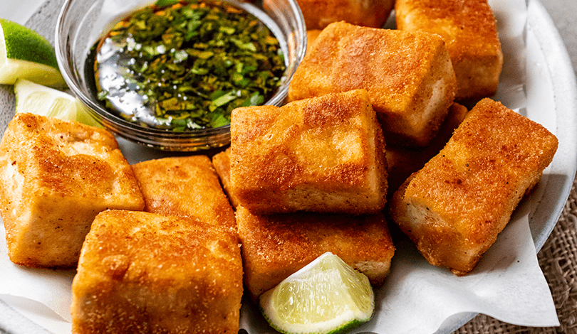 Golden fried tofu cubes served on a plate with lime wedges and a small bowl of dipping sauce with herbs.