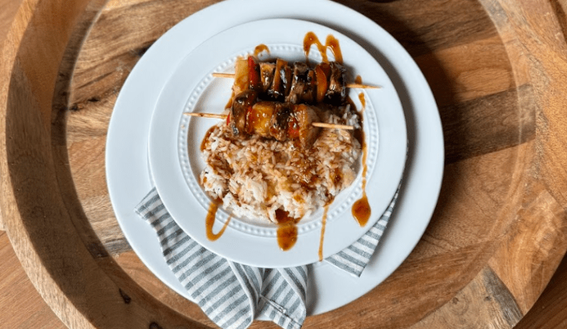 A white plate with grilled meat skewers on a bed of rice, drizzled with brown sauce, set on a striped napkin and wooden tray.