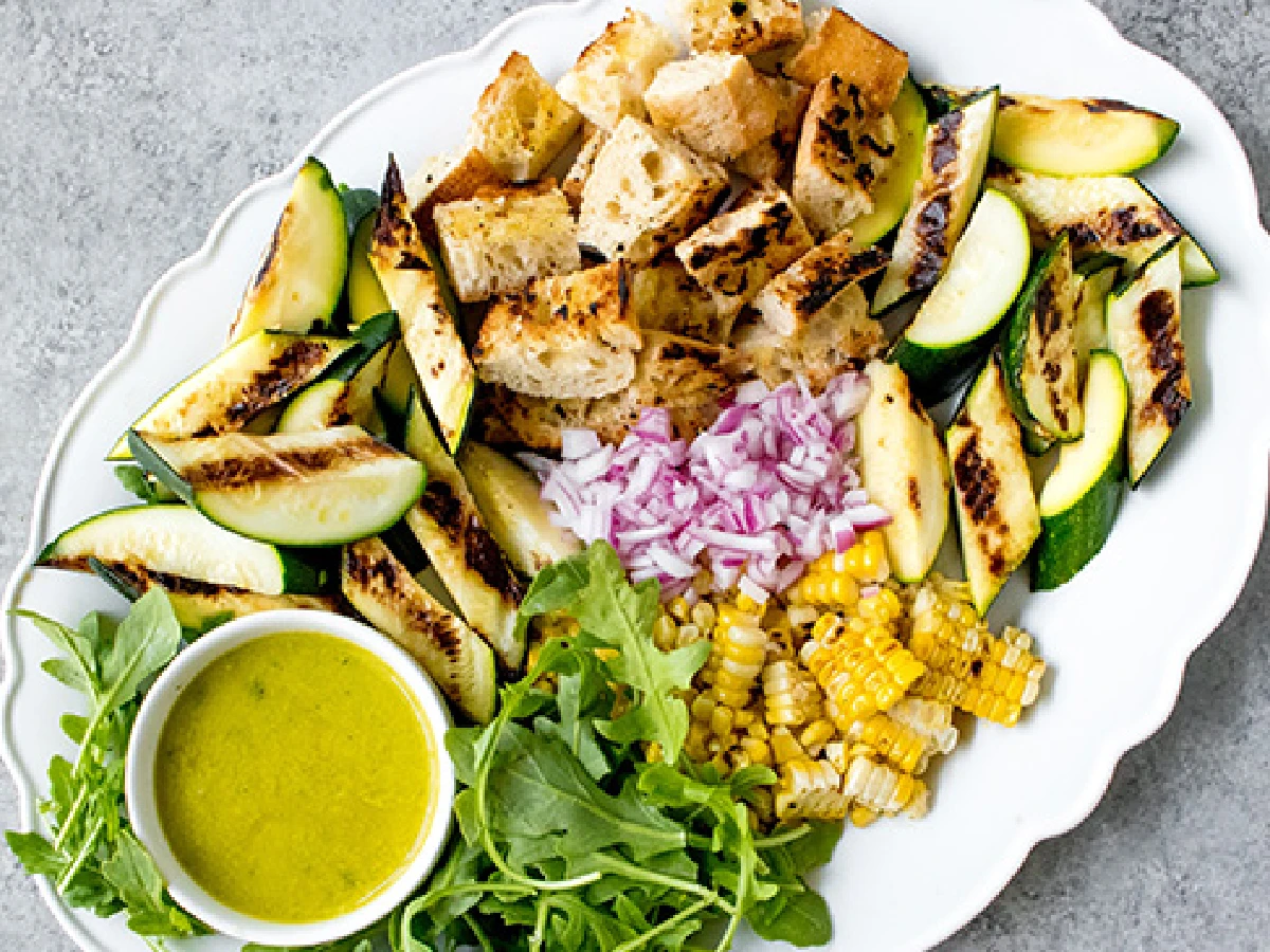 A plate with grilled zucchini, cubed bread, chopped red onion, corn, arugula, and a small bowl of yellow dressing.