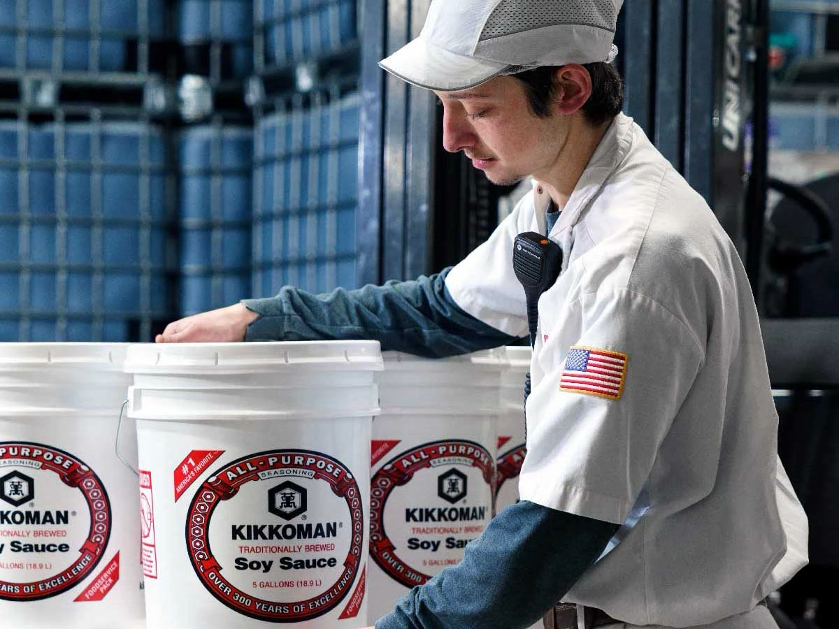 A worker in uniform with an American flag patch handles large white buckets labeled "Kikkoman Soy Sauce" in an industrial setting.