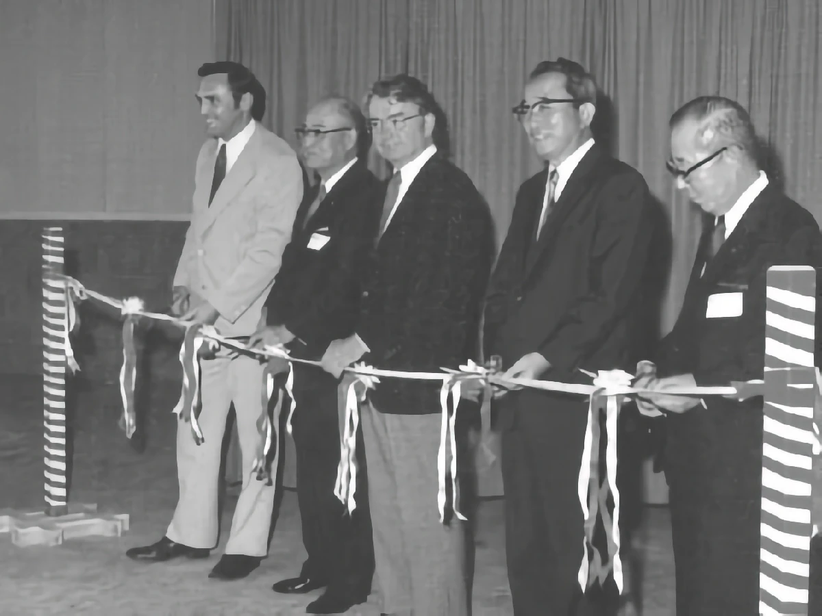 Five men in suits stand in a row holding a ribbon at a ribbon-cutting ceremony, with striped posts on either side and a curtain in the background.
