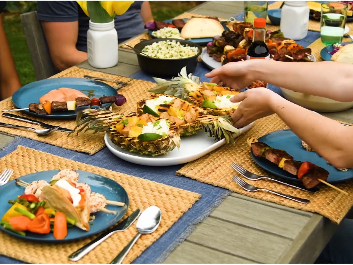 People seated at an outdoor table set with woven placemats, blue plates, pineapple bowls, skewers, salad, bread, and various dishes, with hands serving food.