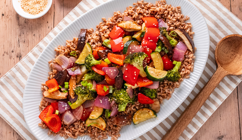 A white oval plate with cooked brown rice topped with sautéed mixed vegetables, including broccoli, red bell peppers, zucchini, onions, and sesame seeds. A wooden spoon is nearby.