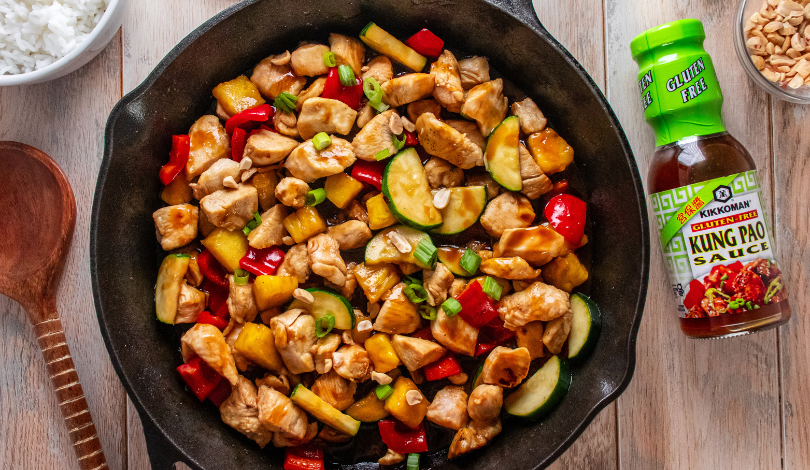A skillet filled with cooked chicken, zucchini, red bell peppers, and pineapple chunks, next to a bottle of Kikkoman Kung Pao sauce, peanuts, and a bowl of white rice.