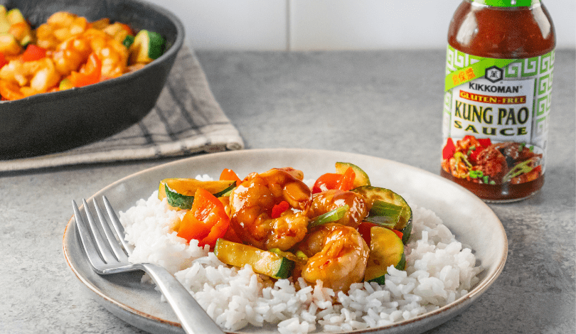 A plate of white rice topped with stir-fried vegetables and shrimp in sauce, next to a bottle of Kikkoman Kung Pao Sauce.