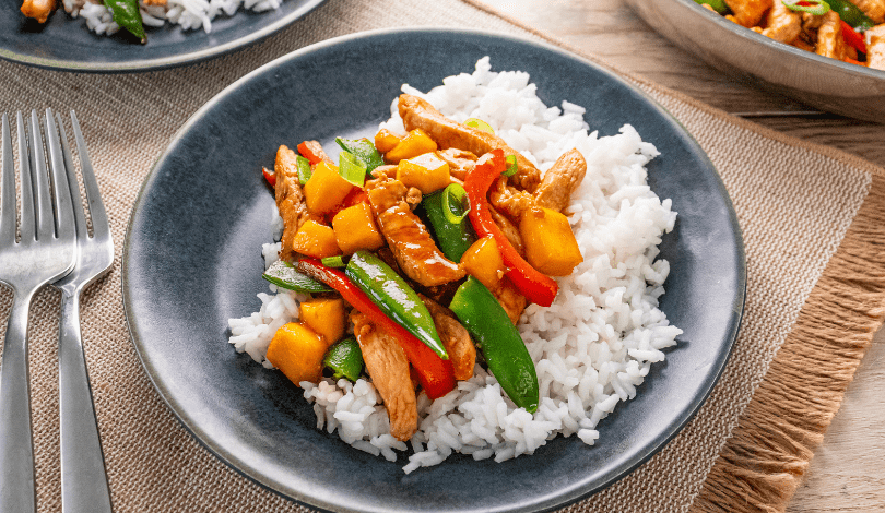A plate of white rice topped with stir-fried chicken, bell peppers, snap peas, and mango pieces, placed on a gray plate with a fork beside it.