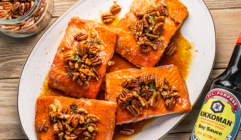Five salmon fillets topped with glazed pecans are arranged on a white oval plate, with a bottle of Kikkoman soy sauce and a jar of pecans nearby.