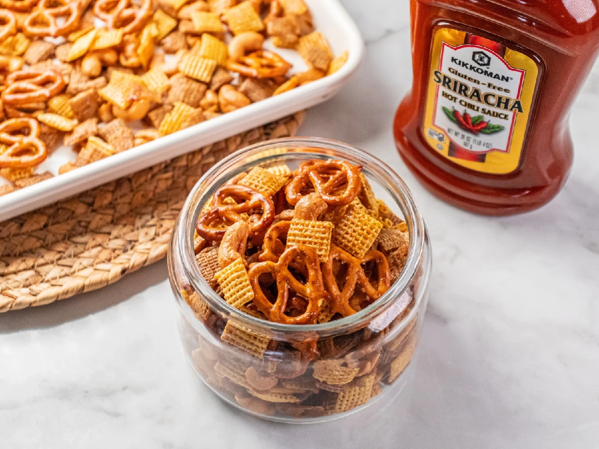 A glass jar filled with snack mix containing pretzels and Chex cereal sits next to a bottle of Kikkoman Sriracha hot chili sauce and a tray of more snack mix.