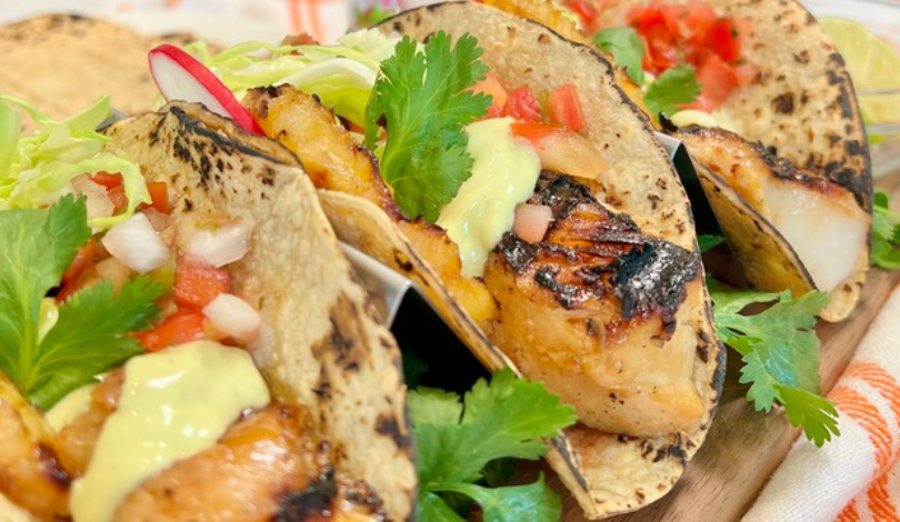 Three grilled fish tacos in charred corn tortillas, topped with diced tomatoes, onions, cilantro, shredded lettuce, and a drizzle of creamy sauce.