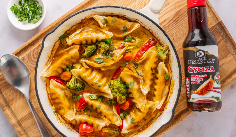 A skillet of dumplings with vegetables in sauce, garnished with herbs, next to a bottle of Kikkoman Gyoza Dipping Sauce on a wooden board.