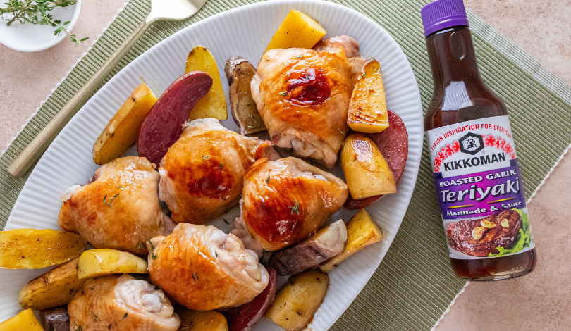 A plate of roasted chicken thighs with potato wedges is beside a bottle of Kikkoman Roasted Garlic Teriyaki Marinade & Sauce.
