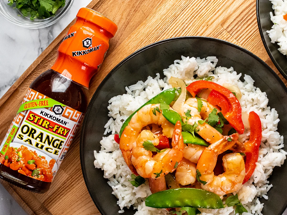 A bowl of white rice topped with shrimp, snap peas, and red bell peppers next to a bottle of Kikkoman stir-fry orange sauce on a wooden surface.