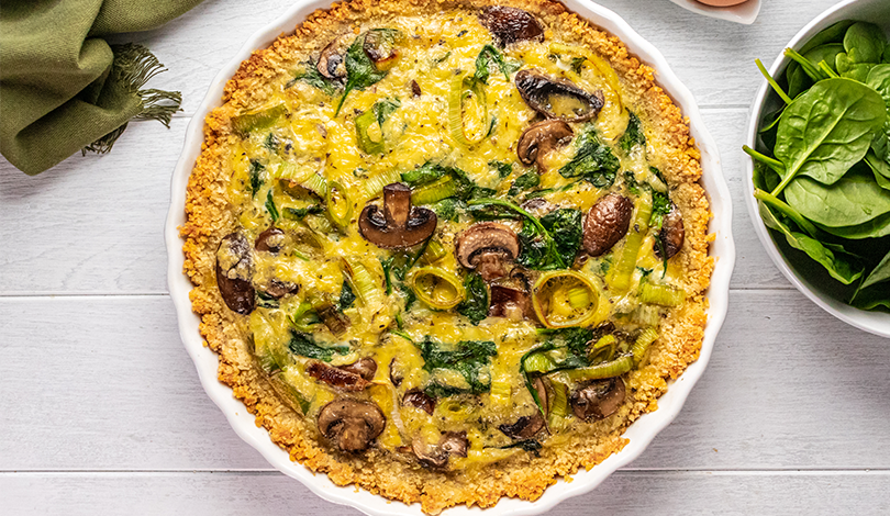 A baked vegetable quiche with mushrooms, spinach, and leeks in a pie dish, shown on a white wooden surface next to a bowl of fresh spinach leaves.