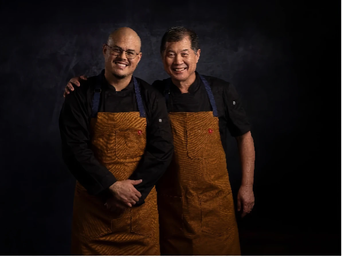 Two men wearing black shirts and brown aprons stand side by side, smiling, with one man resting his arm on the other's shoulder against a dark background.