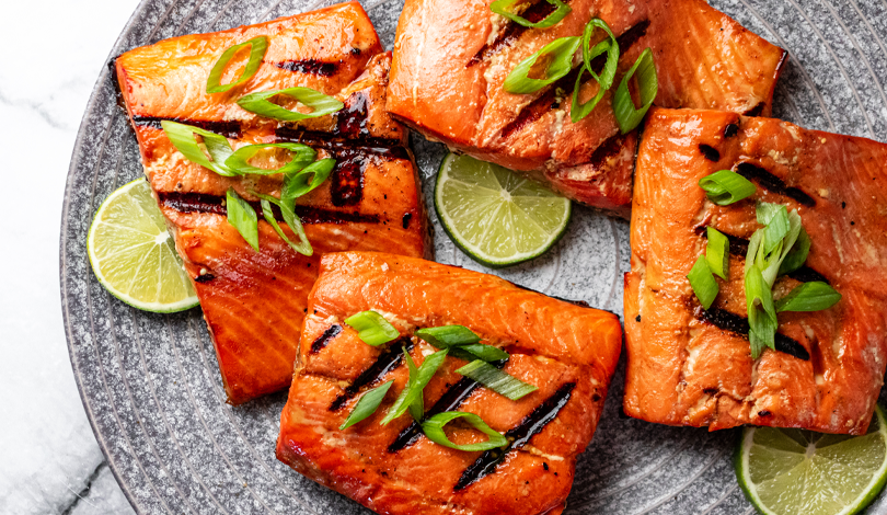 Four grilled salmon fillets topped with sliced green onions are arranged on a gray plate with lime slices.