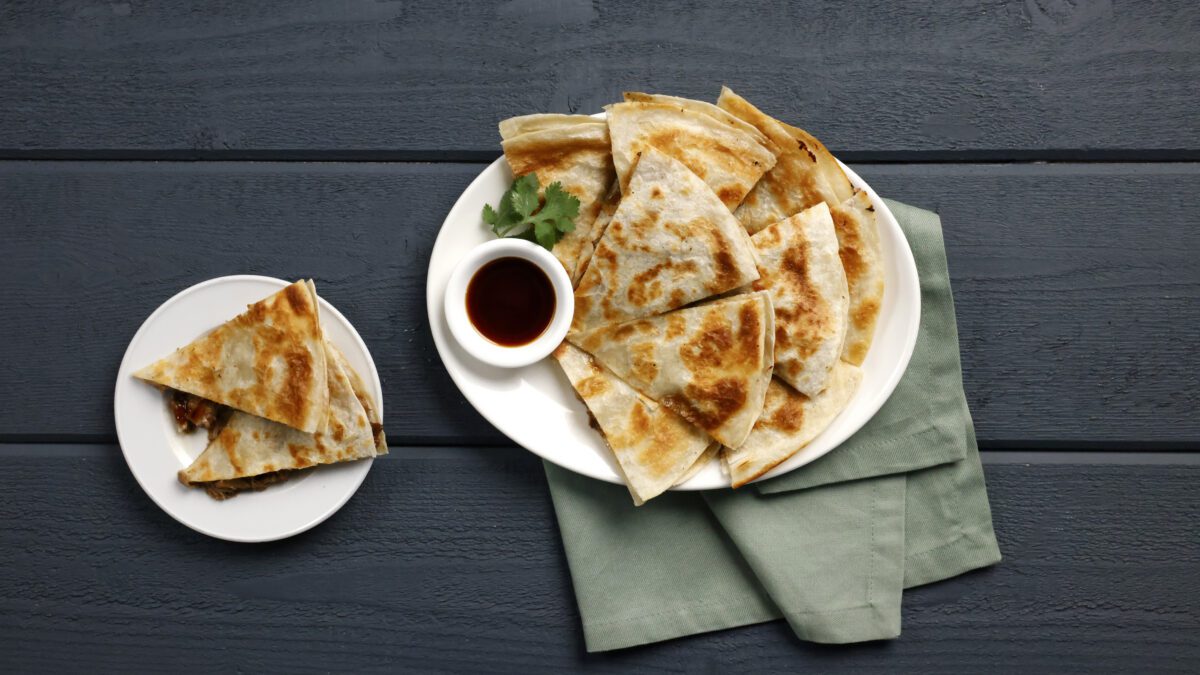 A plate of quesadilla wedges with a small bowl of sauce, garnished with cilantro, next to a smaller plate holding two quesadilla pieces on a dark wooden surface.