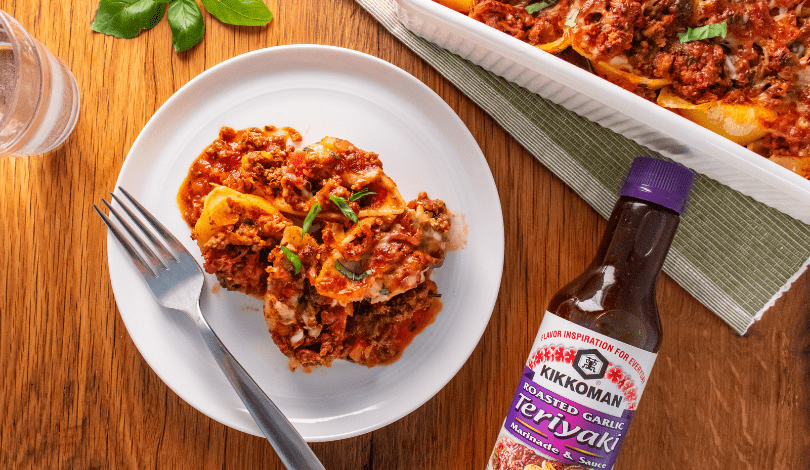 A plate of stuffed pasta shells with meat sauce next to a casserole dish and a bottle of Kikkoman Roasted Garlic Teriyaki Sauce on a wooden table.
