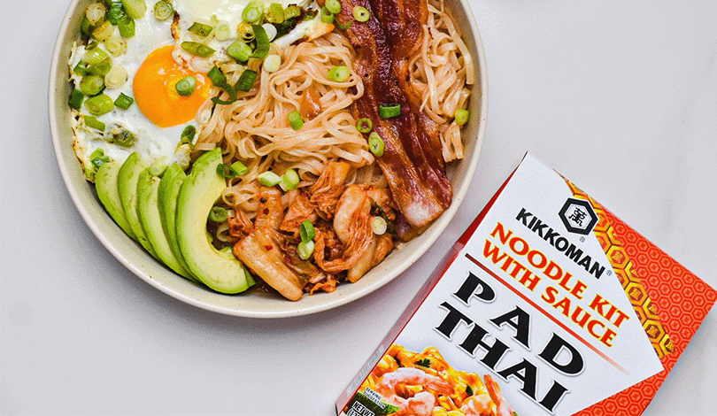 A bowl of Pad Thai noodles with a fried egg, bacon, avocado slices, kimchi, and green onions next to a Kikkoman Pad Thai noodle kit box.