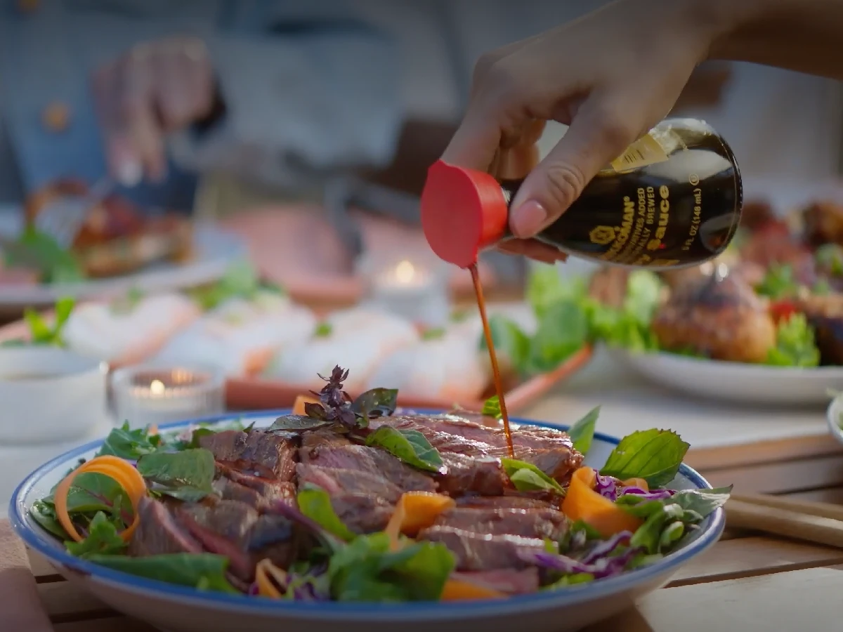 A hand pours soy sauce from a bottle onto a plate of sliced steak with salad garnishes on a table set for a meal.