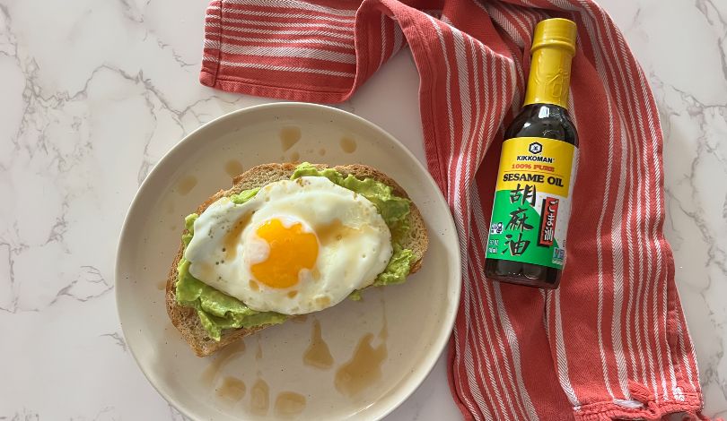 A slice of avocado toast topped with a sunny-side-up egg on a plate, drizzled with sauce, next to a bottle of sesame oil and a red striped kitchen towel on a marble surface.