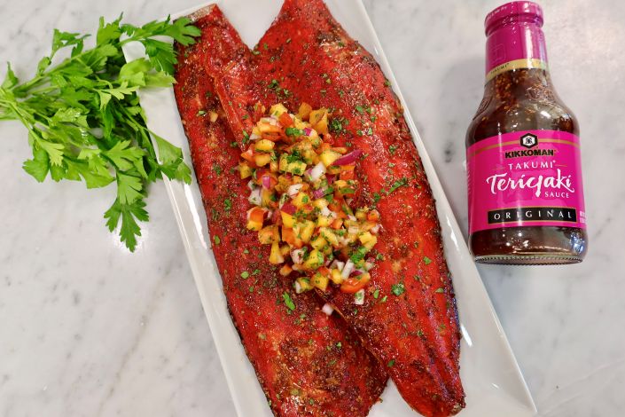 Two grilled salmon fillets topped with a peach salsa are served on a white plate next to a bunch of parsley and a bottle of Kikkoman Takumi Teriyaki Sauce.