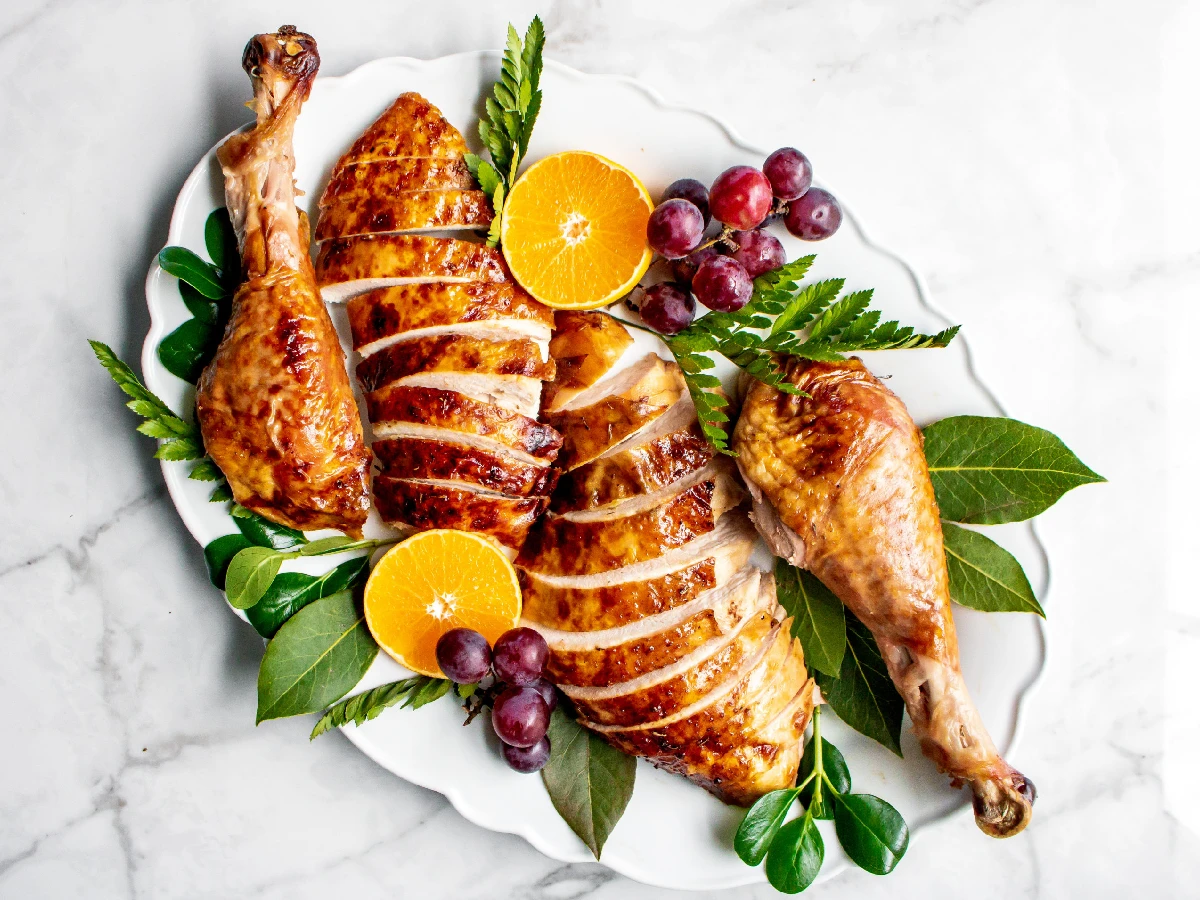 Roasted turkey breast and drumsticks sliced on a white platter, garnished with orange halves, grapes, and green leaves.