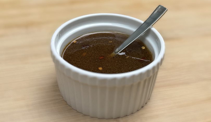 A small white ramekin filled with brown liquid sauce, with visible chili flakes and a metal spoon resting inside.
