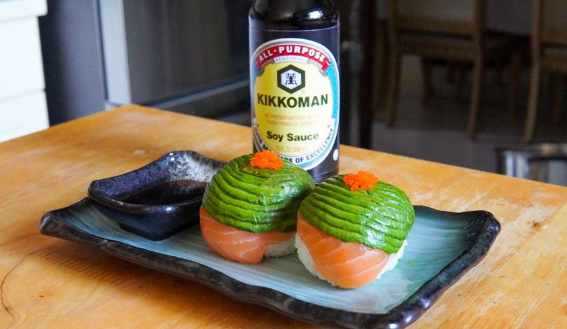 Two pieces of sushi with avocado and salmon, topped with orange roe, are served on a rectangular plate with a soy sauce dish and a bottle of Kikkoman soy sauce in the background.