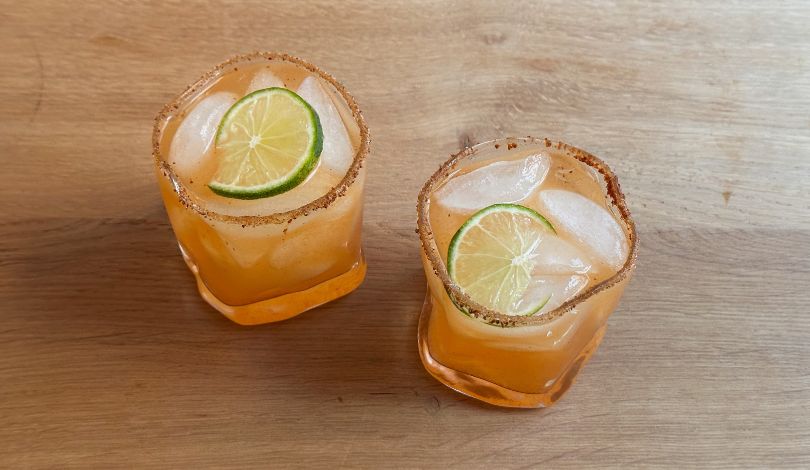 Two glasses of an orange-colored drink with ice, lime slices, and chili powder rims sit on a wooden surface.
