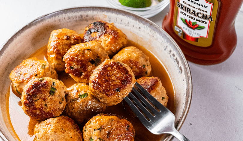 A plate of browned meatballs in sauce with a fork, next to a bottle of Sriracha hot chili sauce and a bowl of limes.