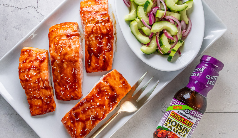 Four pieces of glazed salmon on a white plate, served with a side of cucumber and red onion salad and a bottle of gluten-free hoisin sauce.