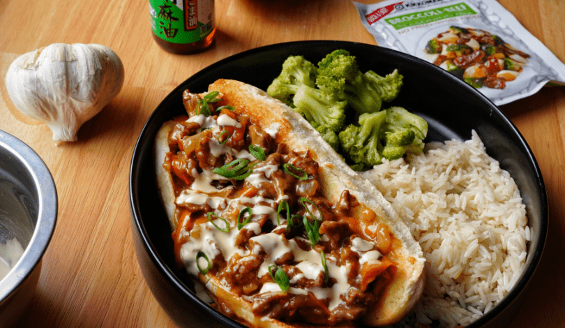 A plate with a beef sandwich topped with sauce and green onions, served with steamed broccoli and white rice. Garlic, a sauce bottle, and a broccoli beef packet are nearby.