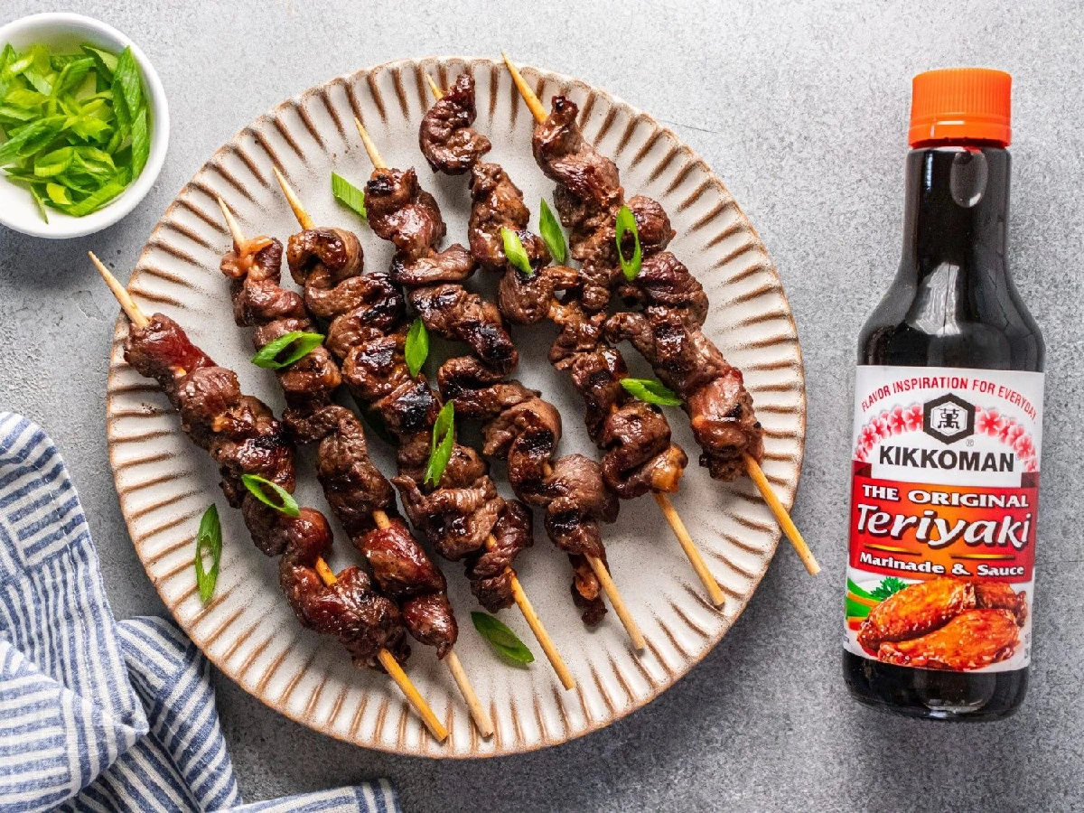 A plate of grilled beef skewers garnished with green onions, next to a bottle of Kikkoman Teriyaki marinade and sauce, with a small bowl of chopped green onions.